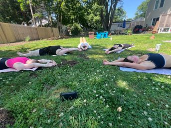 Group of five people doing outdoor yoga on mats, lying with arms stretched overhead in a sunny suburban backyard with trees, wooden fence, and houses