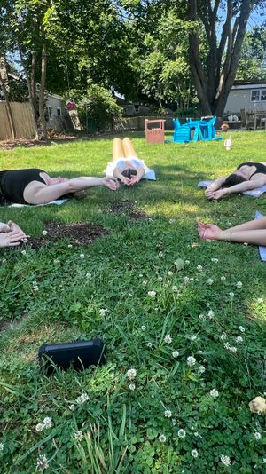 Group outdoor yoga in a sunny suburban backyard — participants lying on mats on a grassy lawn, stretching arms overhead with trees, wooden fence and patio in the background.