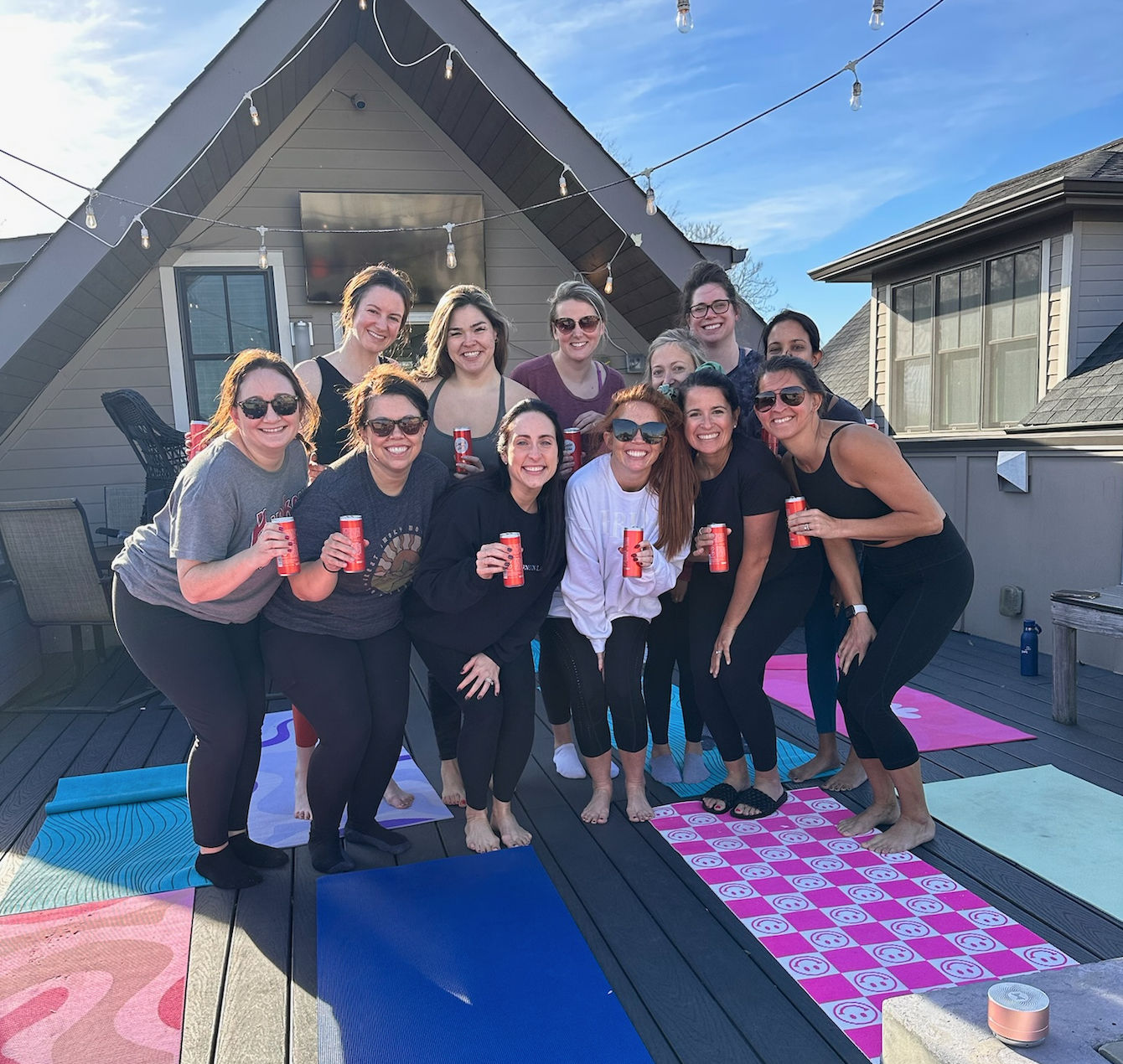 Smiling group of women on a sunny rooftop deck with yoga mats and string lights, posing together and holding canned drinks in front of an A-frame roof.