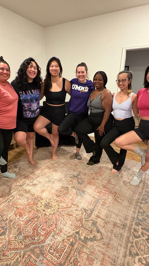 Seven smiling women in workout clothes doing tree pose in a cozy living room during a home yoga session on a large patterned rug.