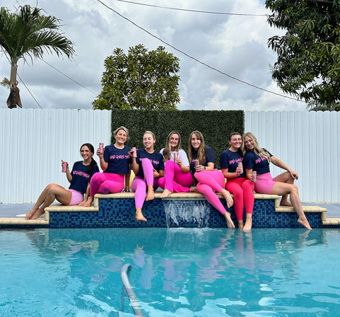 Seven women in matching navy tees and bright pink leggings sitting on a backyard pool ledge, laughing and holding drinks with palm trees and a cloudy sky in the background.