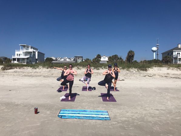 Group of five people practicing beach yoga in tree pose on purple mats on a sunny sandy shoreline with coastal houses and a water tower under a clear blue sky
