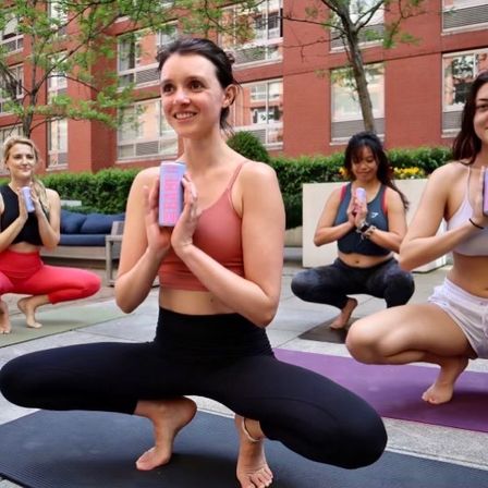 Smiling women in an outdoor urban courtyard yoga class, balancing in a low squat on mats while holding pink yoga blocks at chest — city fitness session