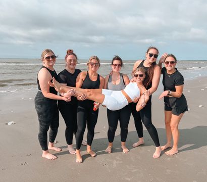 Seven smiling women in activewear and sunglasses on a sandy oceanfront beach; six friends lift one friend horizontally with gentle waves and a cloudy sky behind them.