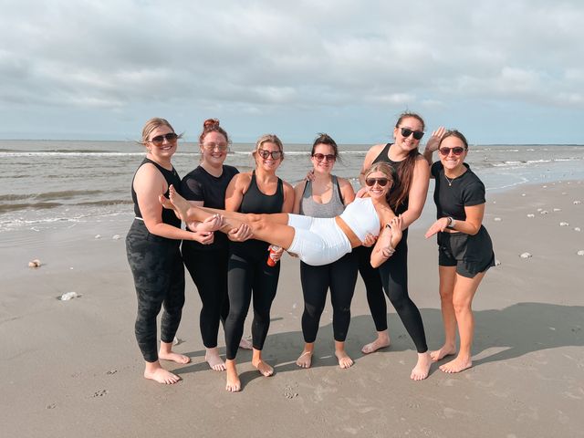 Seven smiling women in activewear and sunglasses on a sandy oceanfront beach; six friends lift one friend horizontally with gentle waves and a cloudy sky behind them.