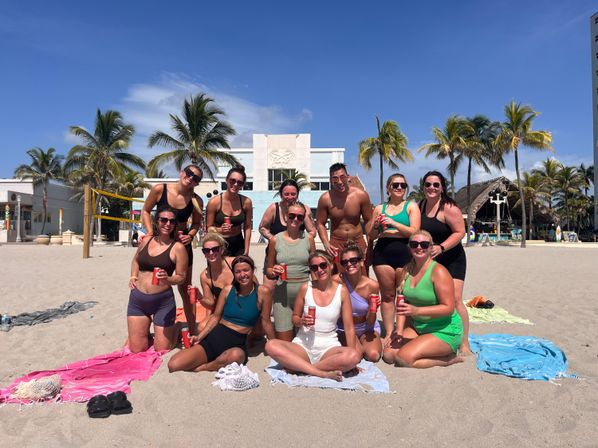 Large group of people in swimsuits posing on a sunny tropical beach at a palm-lined beachfront resort, towels on the sand and drinks in hand near a beach volleyball net.