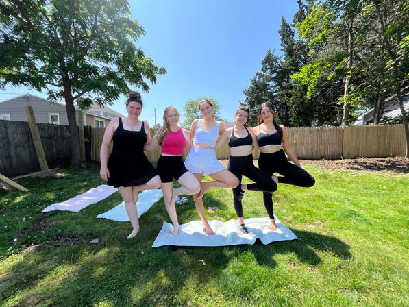 Five friends doing yoga tree pose on mats in a sunny suburban backyard with a wooden fence and tall trees