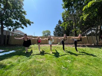 Sunny suburban backyard yoga: five people balancing in Warrior III pose in a row, holding hands above a green lawn with a wooden fence and trees in the background.