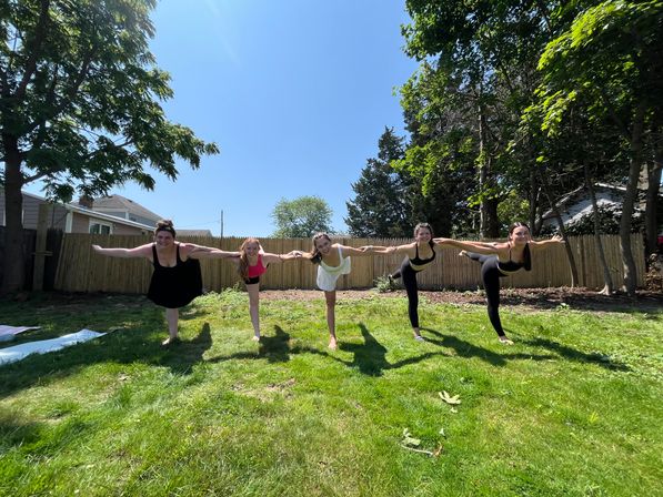 Sunny suburban backyard yoga: five people balancing in Warrior III pose in a row, holding hands above a green lawn with a wooden fence and trees in the background.