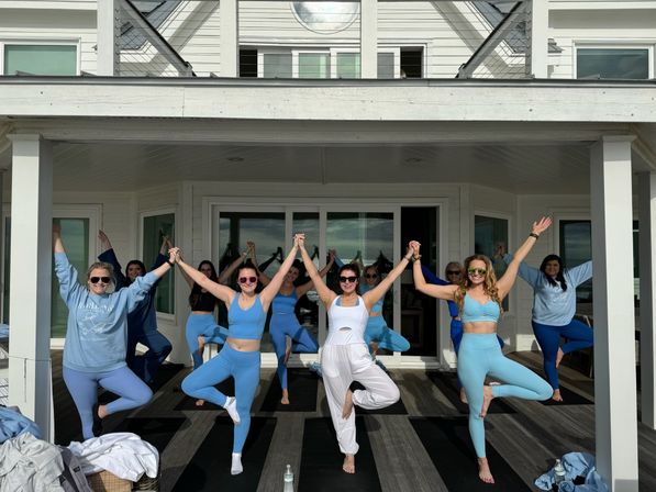 Group of women in blue activewear holding hands and balancing in tree pose on the covered deck of a white coastal-style house — sunny outdoor group yoga session.