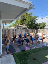 Outdoor backyard yoga class on a sunny patio — a group of participants on mats kneeling and reaching one arm up in a twisting stretch beside a wooden fence and palm trees.