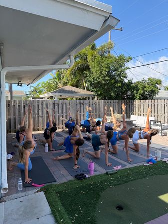Outdoor backyard yoga class on a sunny patio — a group of participants on mats kneeling and reaching one arm up in a twisting stretch beside a wooden fence and palm trees.