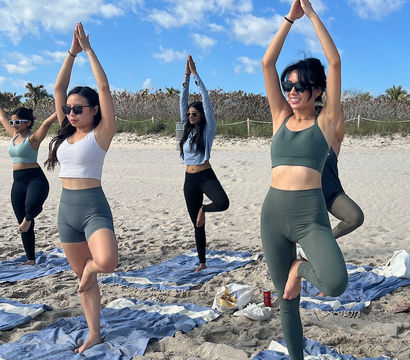Group practicing tree pose on striped beach towels during a sunny outdoor beach yoga session in activewear under a bright blue, partly cloudy sky with sand and coastal vegetation in the background.