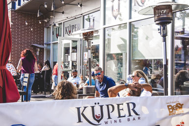 Sunny urban wine‑bar patio with groups chatting and sipping drinks under string lights and tall patio heaters beside a reflective glass storefront.