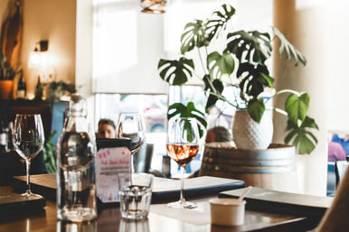 Sunlit cozy cafe bistro table with a glass of rosé, empty wine glasses, water bottle and menus on a wooden surface, large potted monstera plant and barrel accent in the background