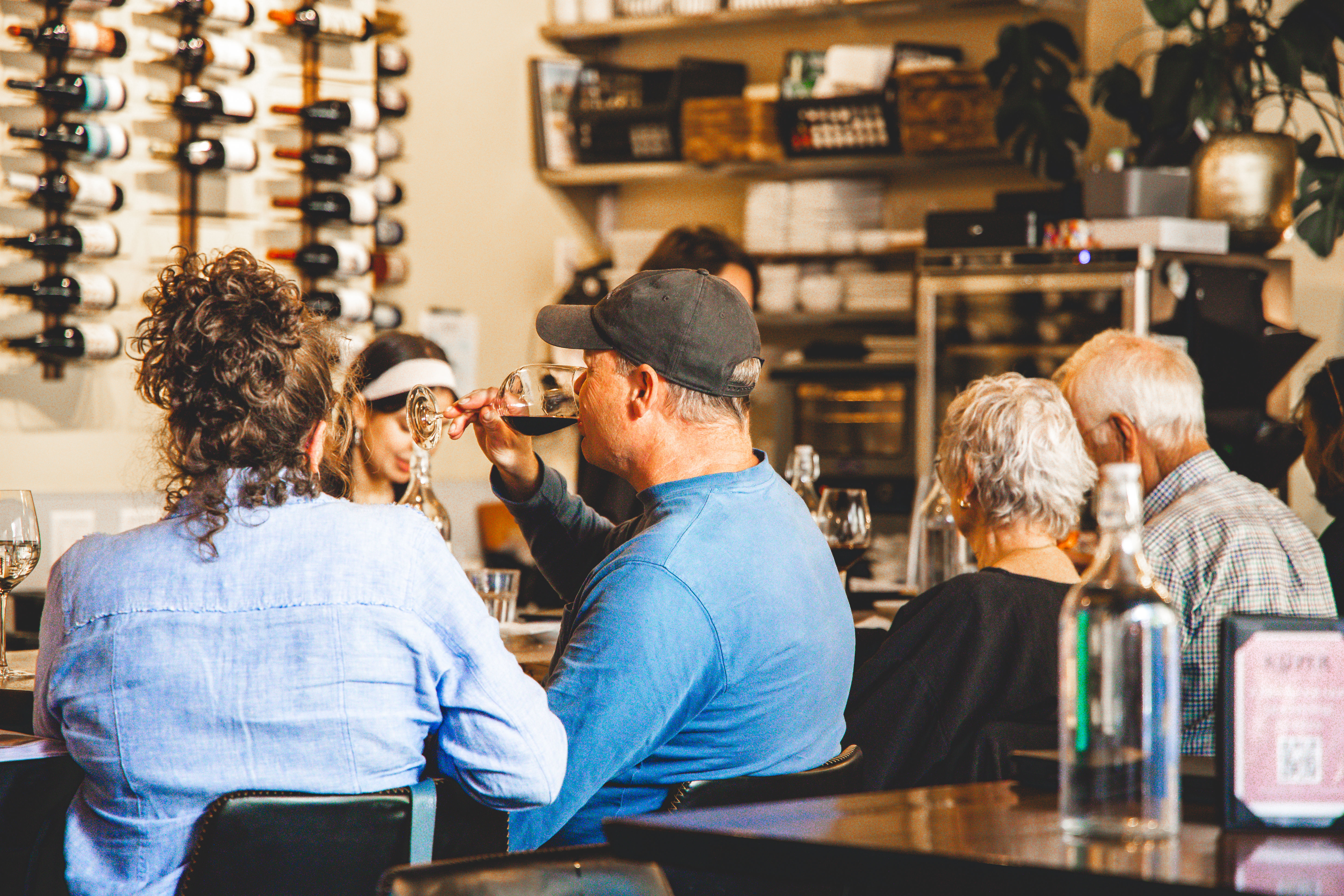 Patrons enjoying wine at a cozy wine bar — a man in a baseball cap sips red wine at a table with a wall-mounted wine rack and shelves in the background.