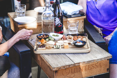 Rustic outdoor patio table with a charcuterie board of cheeses, crackers, olives, pickles, nuts and cured meats, plus a wine bottle and water glass while two people sit nearby