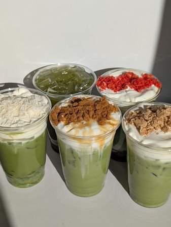 Six iced matcha lattes in clear cups on a white surface, topped with creamy foam and assorted crunchy toppings like shredded coconut, caramel crumbs, red velvet crumbs and brown sugar — bright cafe-style drink spread