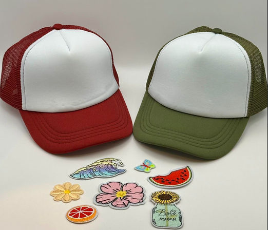 Two blank trucker hats (red and olive) with white fronts arranged beside colorful iron-on patches — wave, butterfly, watermelon, pink flower, daisy, citrus slice and sunflower in a mason jar — for DIY hat customization.