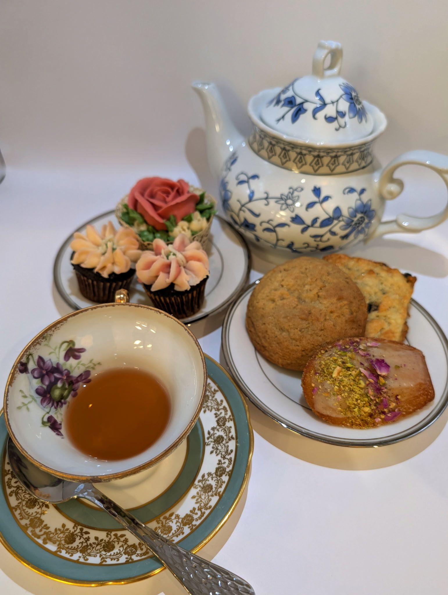 Vintage blue-and-white porcelain teapot and floral china teacup with amber tea, plated scone and iced pistachio cookie plus rose and flower-frosted mini cupcakes on a white backdrop — elegant afternoon tea spread.