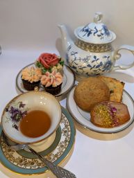 Vintage blue-and-white porcelain teapot and floral china teacup with amber tea, plated scone and iced pistachio cookie plus rose and flower-frosted mini cupcakes on a white backdrop — elegant afternoon tea spread.