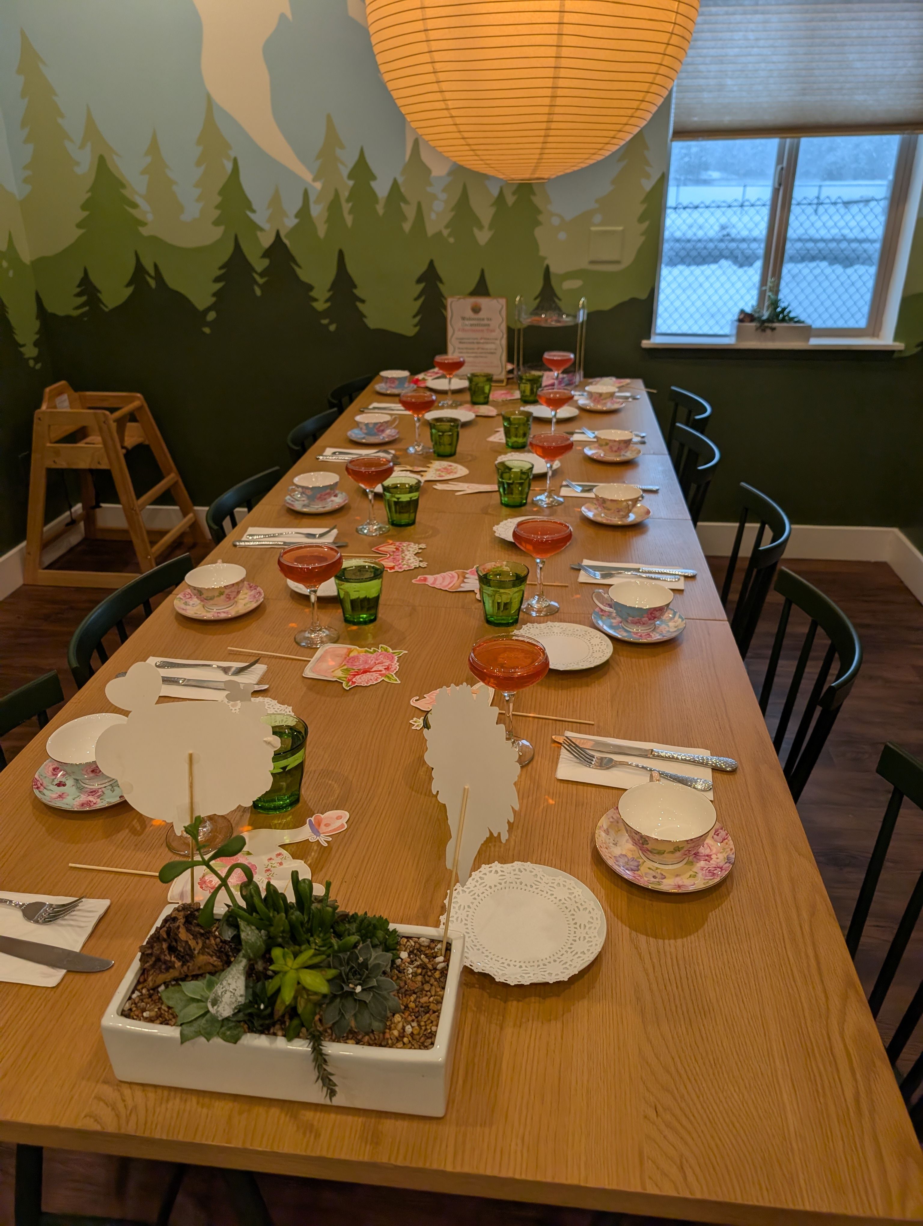 Cozy indoor tea-party setup on a long wooden table with floral teacups, green glasses and pink cocktail coupes, succulent centerpiece and paper place cards under a hanging paper lantern with a forest mural and snowy window.