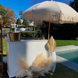 Sunny poolside backyard bar setup with a fringed cream umbrella, white bar counter, gold ice bucket and brass accents, dried pampas grass and palm fan in a white vase beside a blue swimming pool.