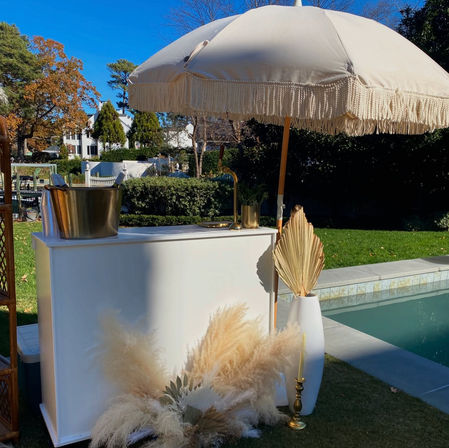 Sunny poolside backyard bar setup with a fringed cream umbrella, white bar counter, gold ice bucket and brass accents, dried pampas grass and palm fan in a white vase beside a blue swimming pool.