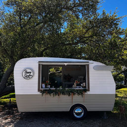 White vintage camper converted into a mobile bar with open serving window, greenery garland and retro hubcap, parked under large oak trees on a sunny day