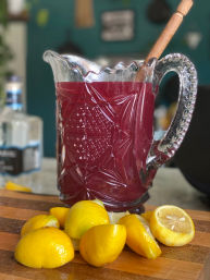 Cut-glass pitcher of deep-red fruit punch with a wooden stirrer and fresh lemon wedges on a wooden cutting board, ready to serve.