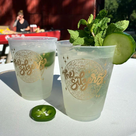 Two frosty summer cocktails in plastic cups with a gold disco design, garnished with cucumber slice, fresh mint and jalapeño slice on a white table at an outdoor event.