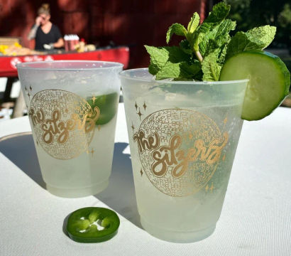 Two frosty summer cocktails in plastic cups with a gold disco design, garnished with cucumber slice, fresh mint and jalapeño slice on a white table at an outdoor event.