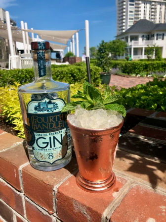 Gin bottle beside a copper julep cup filled with crushed ice and fresh mint on a brick ledge at a sunny coastal garden patio