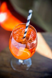 Bright orange Aperol Spritz in a stemmed wine glass with ice, an orange slice and a black-and-white striped straw on a wooden table, top-down view — refreshing summer cocktail.