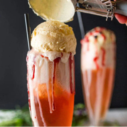 Tall strawberry soda float topped with a creamy vanilla ice cream scoop and red syrup dripping down the glass, black straw and a second blurred float in the background