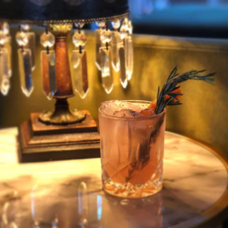 Pink herb-garnished cocktail in a cut-glass rocks glass on a marble table beside a vintage crystal-beaded lamp in a cozy lounge
