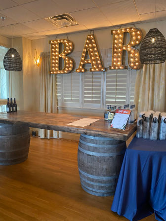 Indoor event bar with large lit marquee "BAR" letters, rustic wooden tabletop supported by wine barrels, wicker pendant lights and shuttered windows.