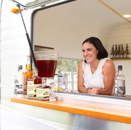 Smiling woman leaning out of a white vintage camper window serving cocktails at a mobile bar, with bottles, glassware and a large iced beverage dispenser on the counter.