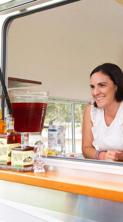 Smiling woman leaning out of a white vintage camper window serving cocktails at a mobile bar, with bottles, glassware and a large iced beverage dispenser on the counter.