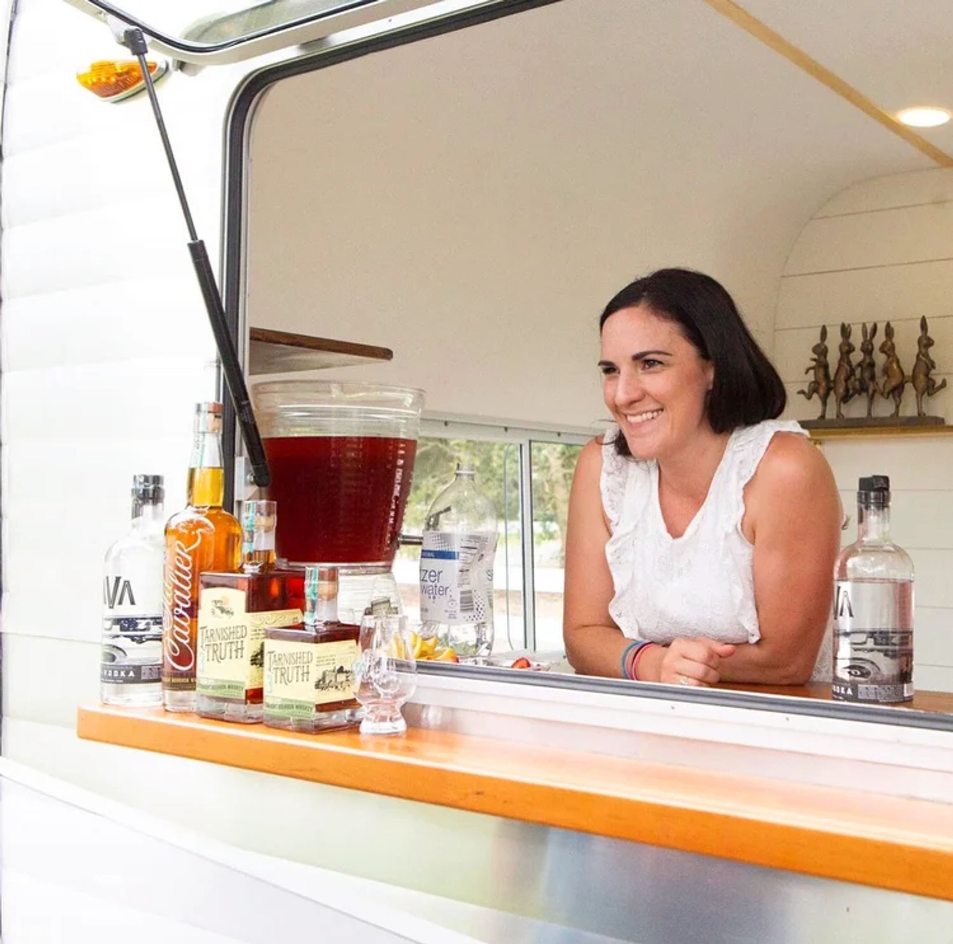 Smiling woman leaning out of a white vintage camper window serving cocktails at a mobile bar, with bottles, glassware and a large iced beverage dispenser on the counter.