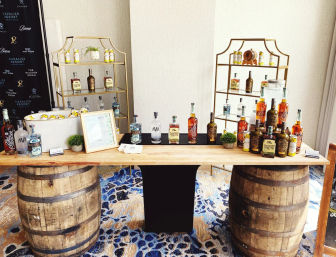 Rustic spirits tasting bar at an indoor event: wooden plank atop two oak barrels displaying whiskey and gin bottles, ice bucket with lemon slices, gold display shelves, framed menu on patterned carpet.
