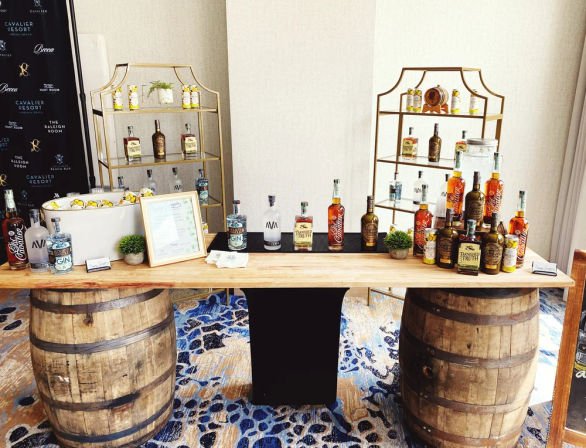 Rustic spirits tasting bar at an indoor event: wooden plank atop two oak barrels displaying whiskey and gin bottles, ice bucket with lemon slices, gold display shelves, framed menu on patterned carpet.