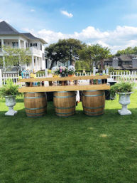 Rustic outdoor bar made from three wooden barrels and a wooden plank on a green lawn, topped with bottles and a colorful floral centerpiece, flanked by stone urn planters with a white picket fence and two‑story house in the background.