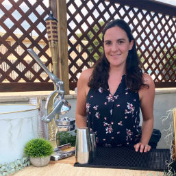 Smiling woman standing behind an outdoor bar on a sunny patio, with a vintage citrus press, metal cocktail shakers, small potted plant and lattice privacy screen with tiki torch in the background.