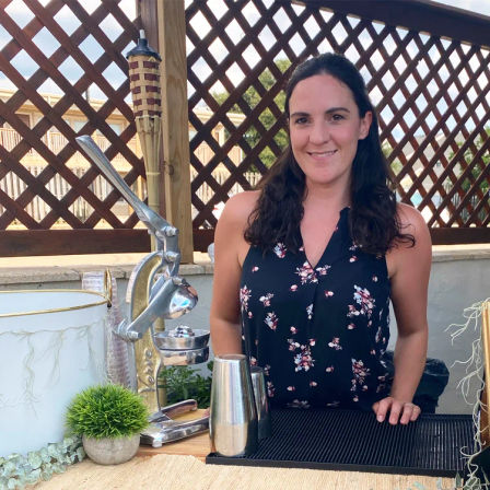 Smiling woman standing behind an outdoor bar on a sunny patio, with a vintage citrus press, metal cocktail shakers, small potted plant and lattice privacy screen with tiki torch in the background.
