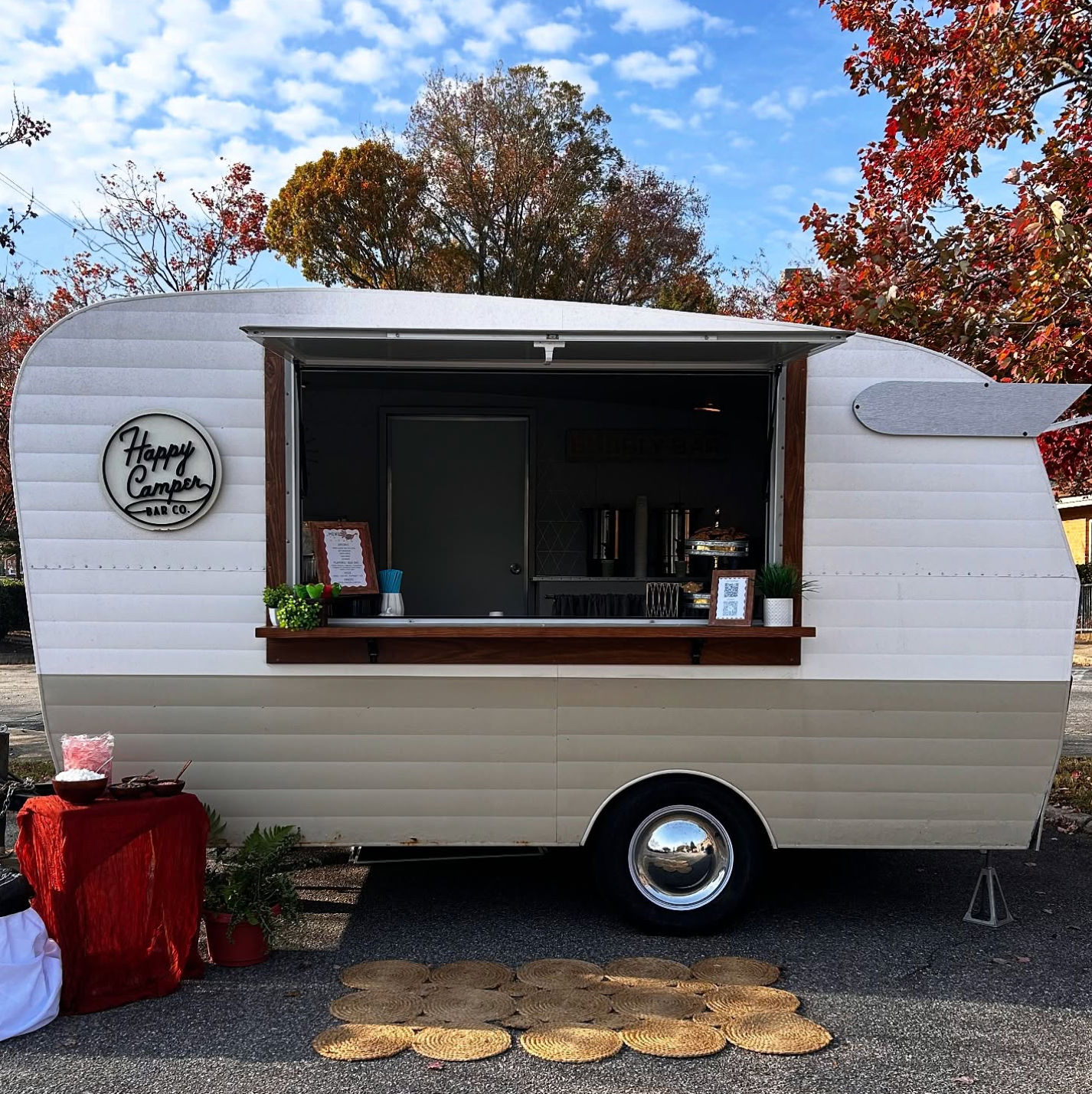 Retro cream-and-beige vintage camper converted into a mobile bar with open serving window, wood counter, small menu sign and potted plants, parked on a leafy autumn street with woven mats in front.