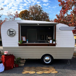 Retro cream-and-beige vintage camper converted into a mobile bar with open serving window, wood counter, small menu sign and potted plants, parked on a leafy autumn street with woven mats in front.