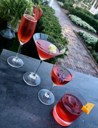 Four rosy cocktails on a dark stone ledge overlooking a garden brick pathway — champagne flute with a strawberry, martini with a white rose, coupe garnished with lavender, and a rocks glass with an orange twist.