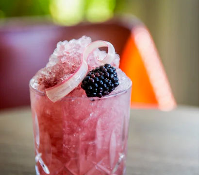 Pink blackberry crushed-ice cocktail in a cut crystal glass, topped with a fresh blackberry and a pale curled rhubarb ribbon, on a wooden table with soft bokeh background