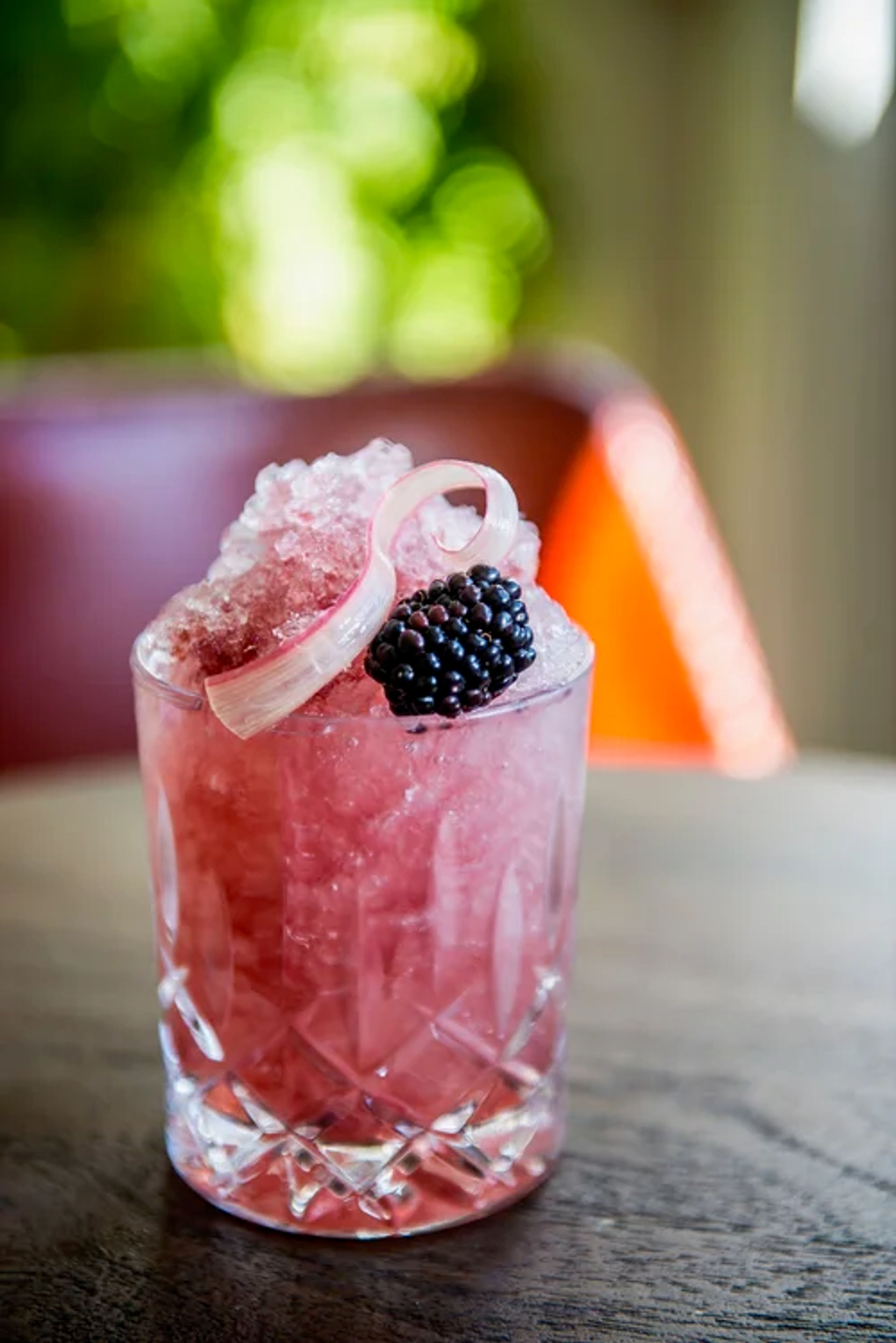 Pink blackberry crushed-ice cocktail in a cut crystal glass, topped with a fresh blackberry and a pale curled rhubarb ribbon, on a wooden table with soft bokeh background